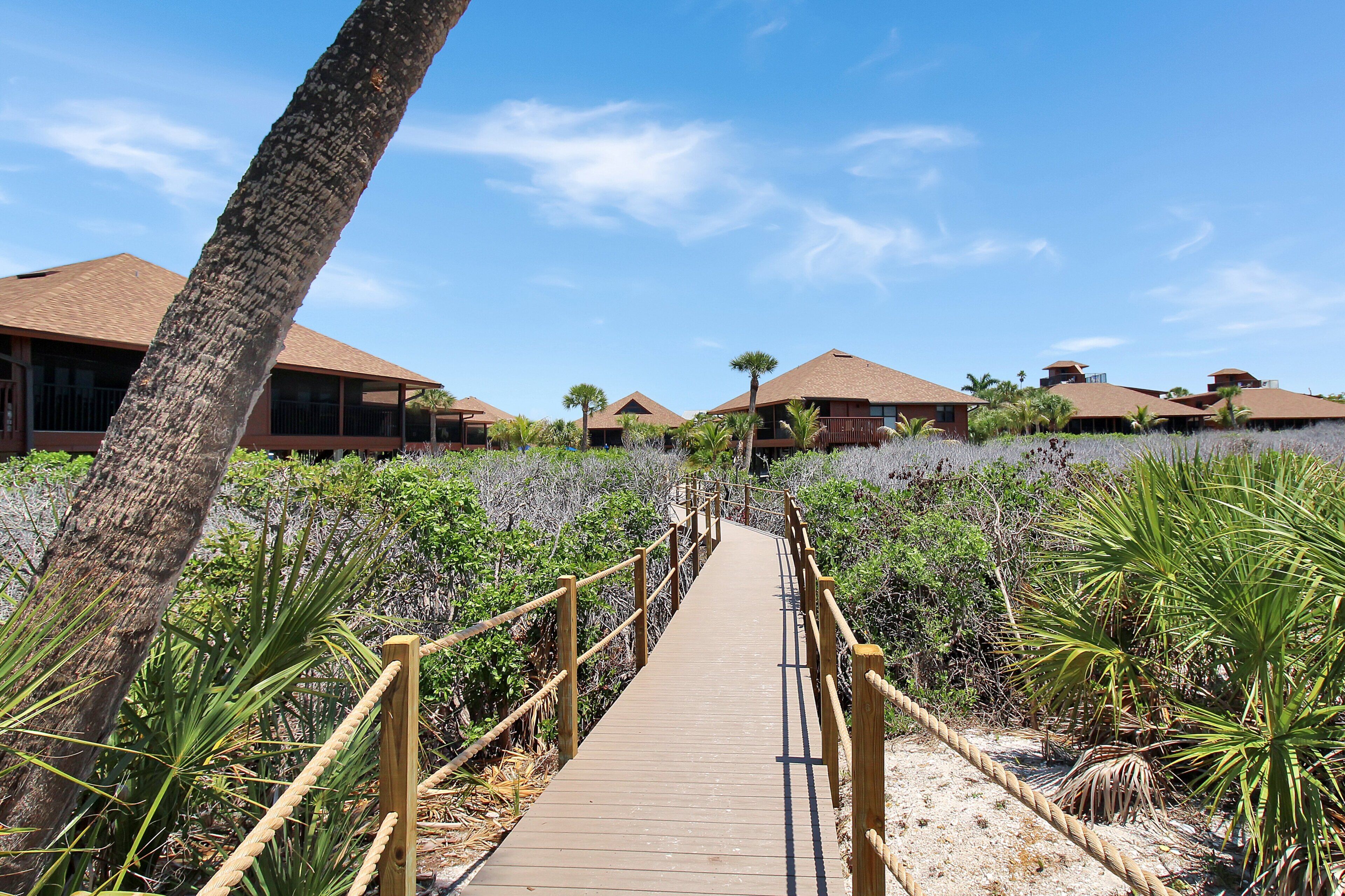 A wooden boardwalk leading to a resort with multiple buildings, surrounded by tropical vegetation.