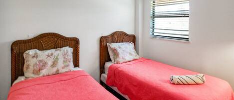 Habitación con dos camas individuales, cada una con cabecera y almohada. Se observa una ventana con persianas en la pared.
