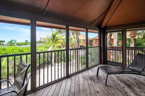 A covered outdoor area with wooden flooring and metal railings, featuring two chairs and a view of a landscaped area with palm trees and a building in the background. - 3BR Villa with rooftop sundeck, gulf views and private beach - pet friendly - Signal Inn 16 (Sanibel)