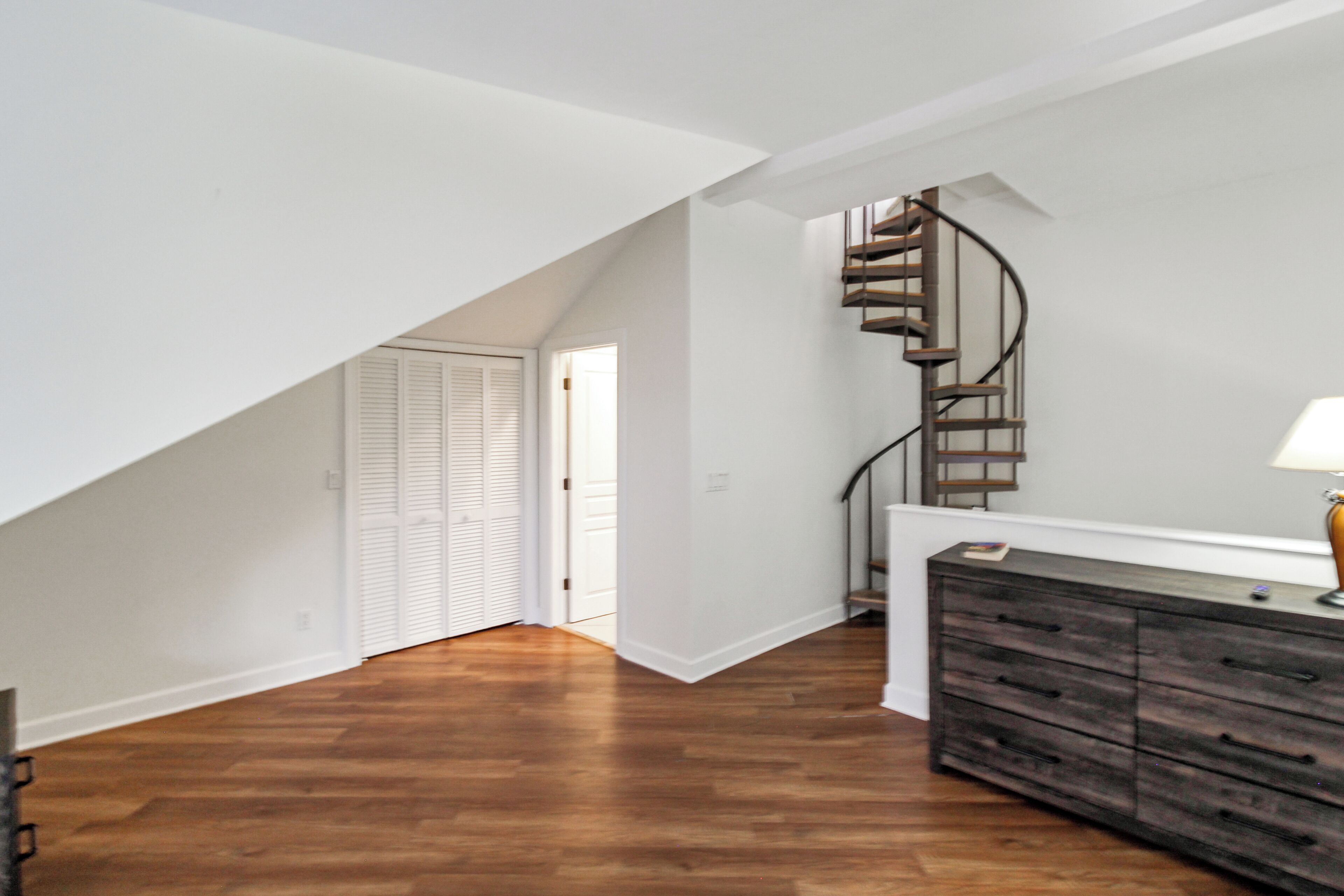 A staircase with a curved design leading to an upper level, a wooden dresser with drawers, and a white door with louvered shutters.
