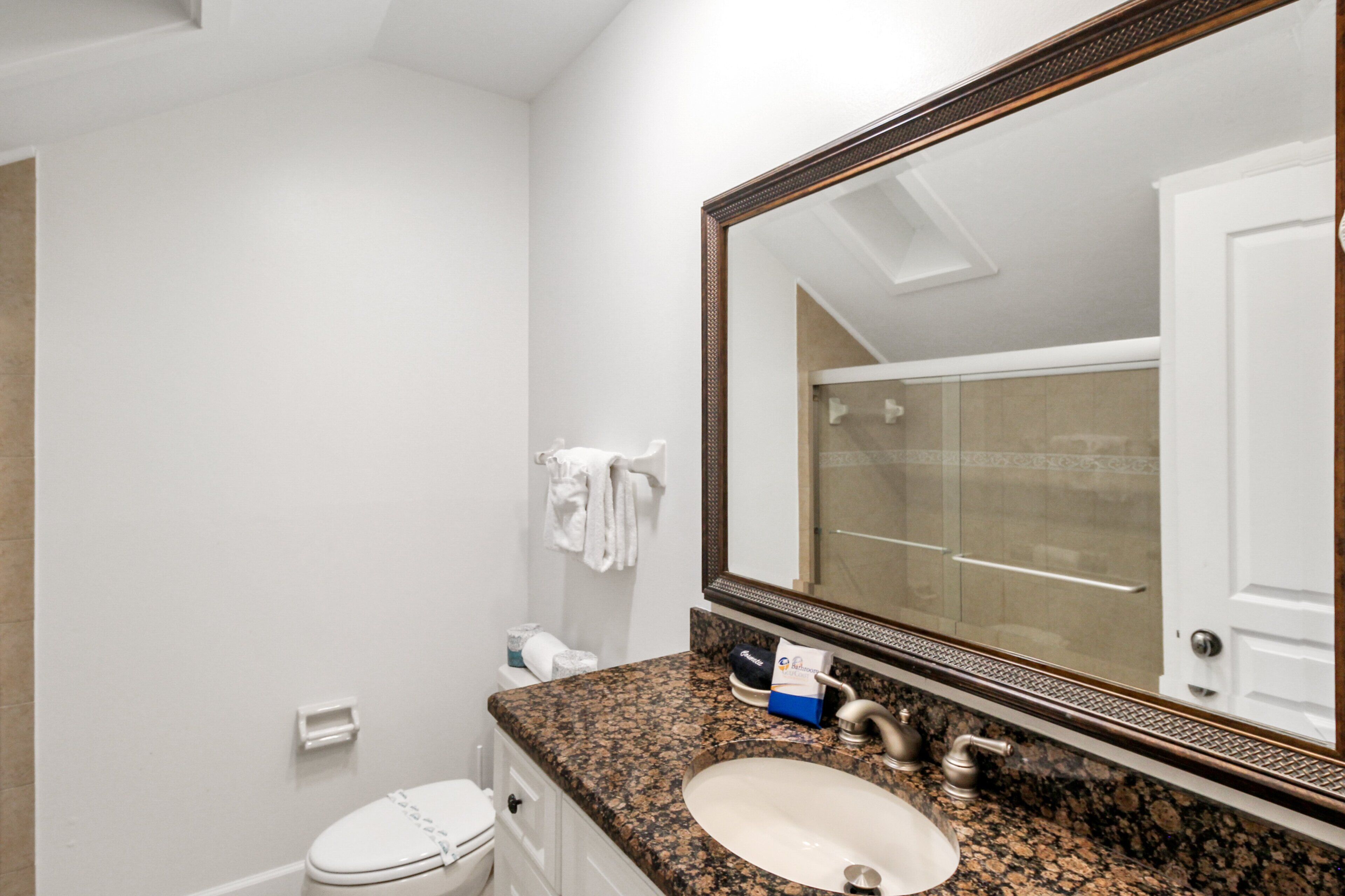 Une salle de bain avec des toilettes blanches, un lavabo doté d’un plan de travail en granit et un grand miroir avec un cadre en bois.