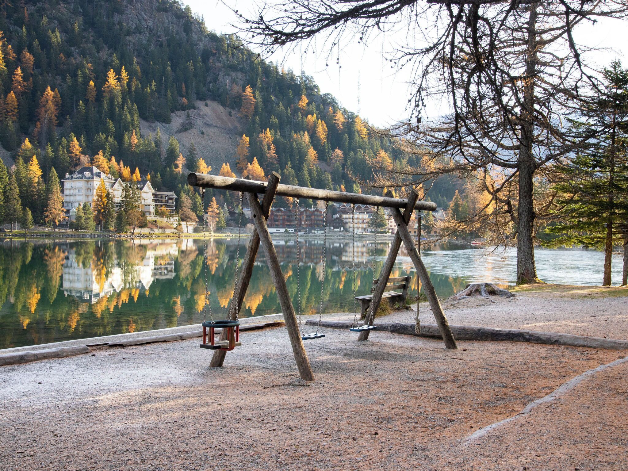 A wooden swing set by a lake with a reflection of buildings and trees in the water.