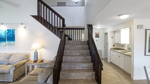 Una escalera con barandillas de madera que lleva a un piso superior, una sala de estar con un sofá y una lámpara, y una zona de cocina con un lavamanos y armarios.