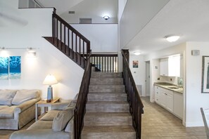 A staircase with wooden railings leading to an upper floor, a living room with a sofa and a lamp, and a kitchen area with a sink and cabinets.