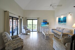 A spacious living room with a sofa, armchair, and a coffee table. There is a television mounted on the wall and a ceiling fan.
