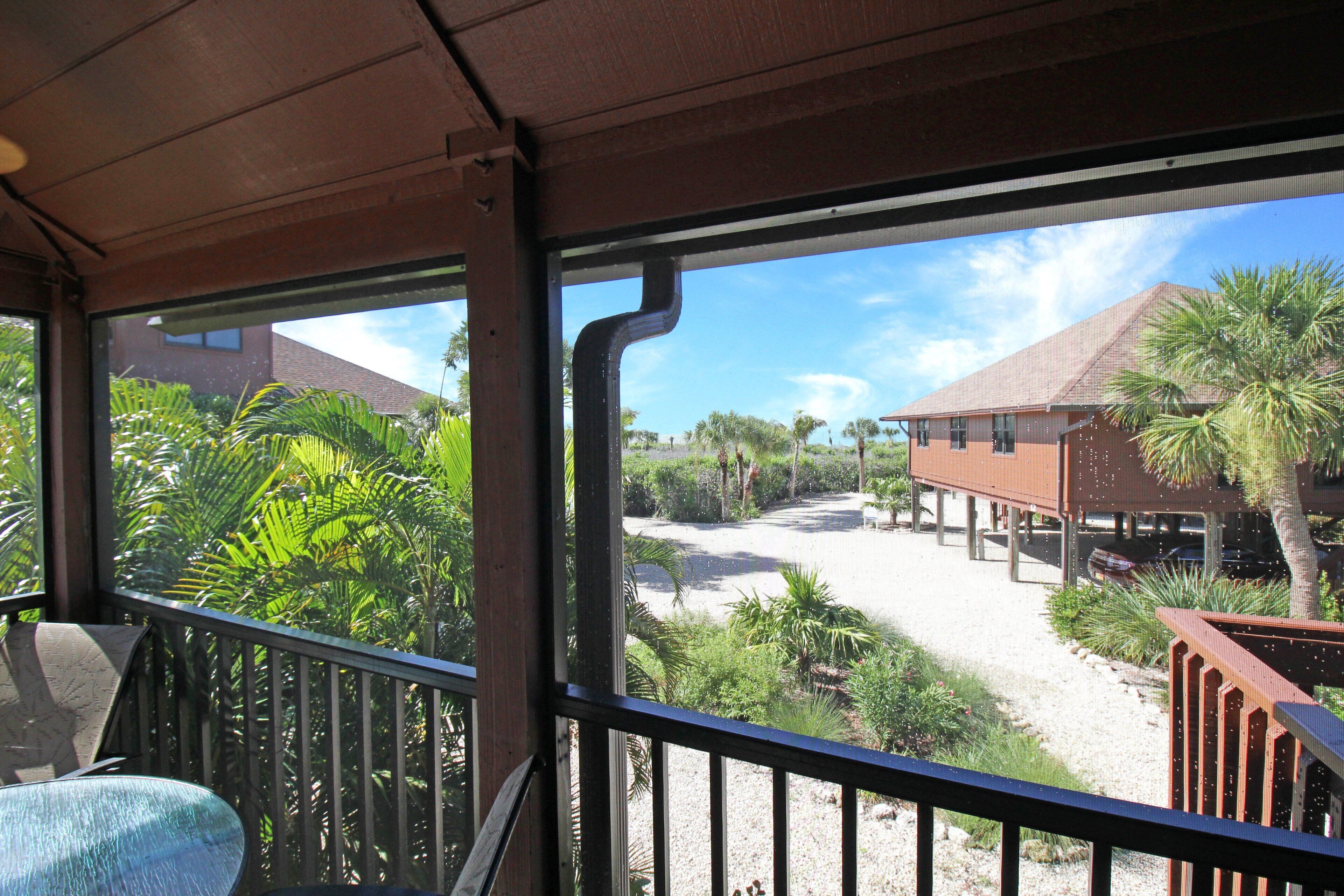 A view from a covered balcony with a railing, overlooking a parking area and buildings with a clear sky.