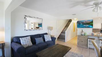 Ein Wohnzimmer mit einer Couch, einem Couchtisch und einem Fernseher. Eine Treppe führt zu einer Küchenzeile.