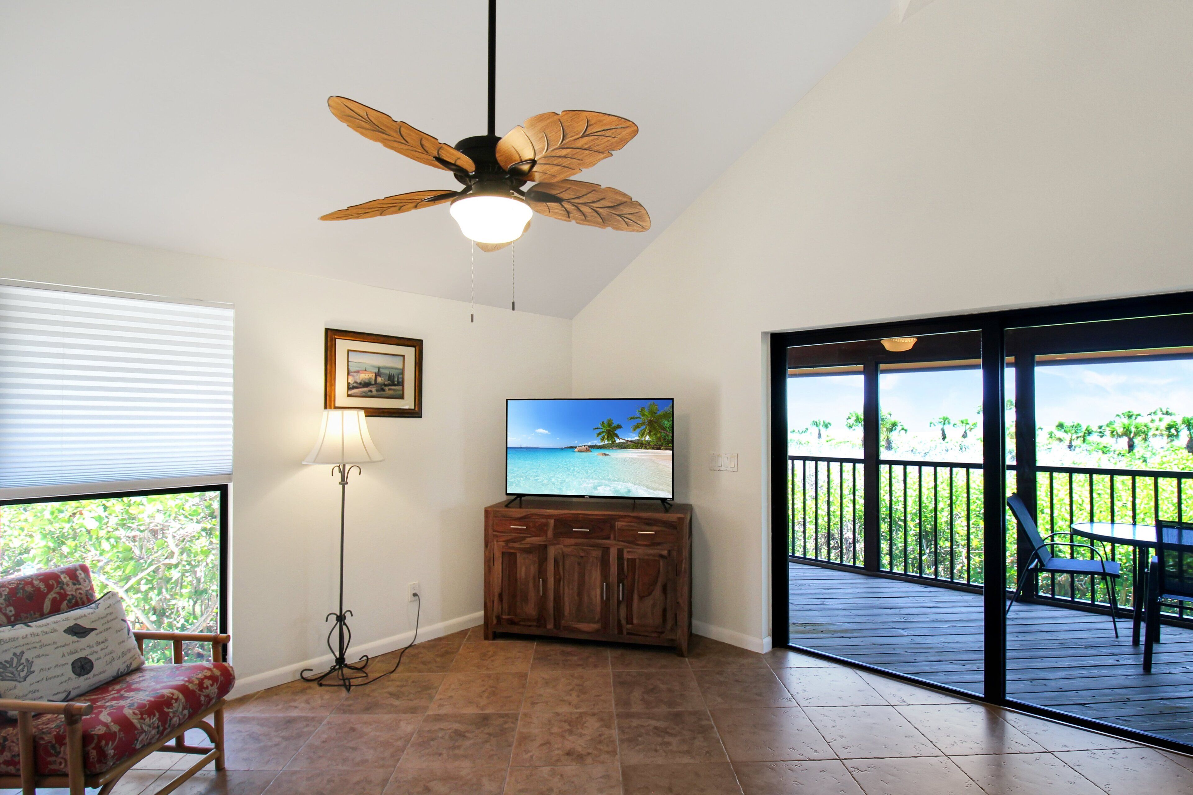 A room with a ceiling fan, a television on a wooden cabinet, a chair, and a view of a balcony with a table and chairs.