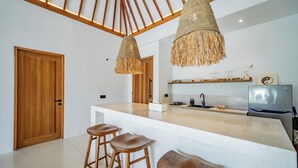 A modern kitchen with a central island, wooden stools, hanging woven light fixtures, and a stainless steel refrigerator. - Villa Rosado Canggu (Canggu)