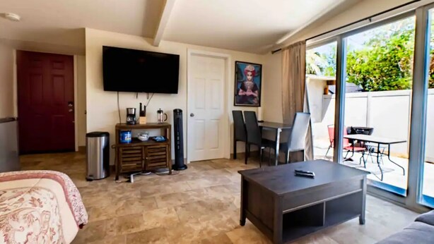 Un salón con televisor de pantalla plana, una mesa de centro y un sofá. Hay una zona de comedor con una mesa y sillas, y una puerta corrediza de vidrio que da a un patio exterior.