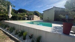 Une piscine bordée d’un muret en pierre, un jardinier rouge et une petite table avec des chaises.