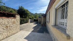 Un bâtiment en pierre avec une terrasse carrelée, une fenêtre ornée de barreaux en métal et une montagne en arrière-plan.