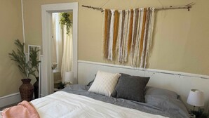Un dormitorio con cama, almohadas, una lámpara de noche, un cuadro en la pared y una planta en maceta.