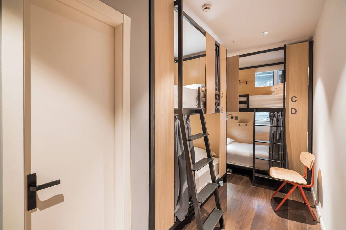A compact hotel room with a bunk bed, a ladder, a chair, and a wardrobe.