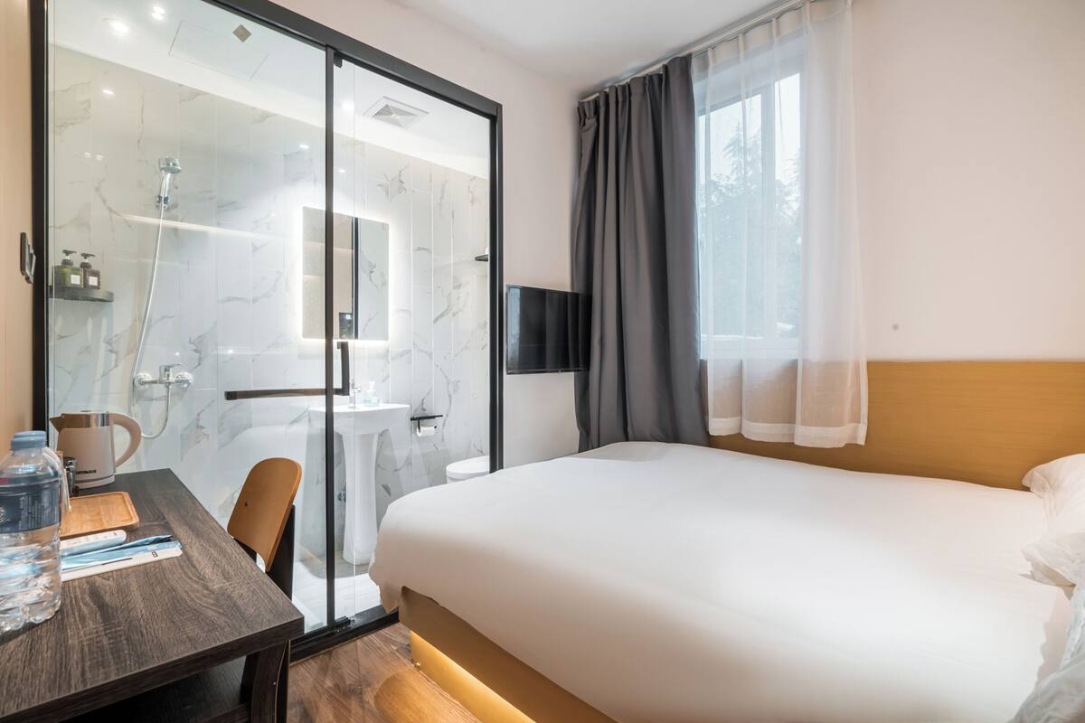 A hotel room with a large bed, a desk with a chair, a shower area, and a TV mounted on the wall.