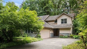 A two-story house with a stone foundation, a covered driveway, and lush greenery surrounding it. - Fantastic Golf and Ski Retreat Surrounded by Trees, Private Hot Tub, Game Room, BBQ (Mount Hood Village)