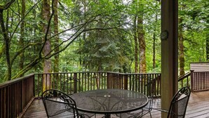 A covered outdoor deck with a round metal table and four chairs, surrounded by a wooden railing, set in a forested area. - Fantastic Golf and Ski Retreat Surrounded by Trees, Private Hot Tub, Game Room, BBQ (Mount Hood Village)