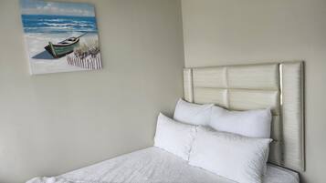 Ein ordentlich bezogenes Bett mit weißen Leinen und einem Kopfteil, ein Gemälde eines Bootes am Strand und eine schlichte Wand.