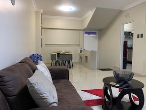 A living room with a brown sofa, a dining table with chairs, a small round table with a bowl, and a kitchen area in the background.