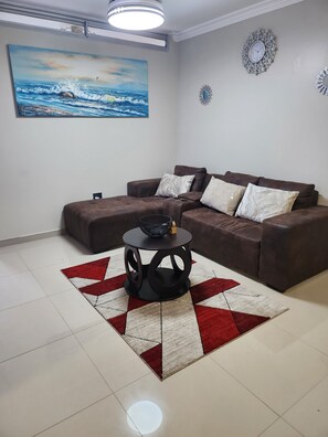 A living room with a brown sectional sofa, a round coffee table with a black base, a red and white patterned rug, a painting of a seascape, and two wall clocks.