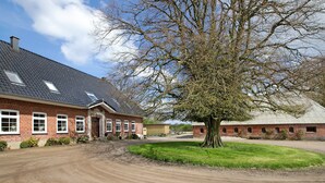 A brick building with a sloped roof, a large tree in front, and a dirt driveway. - Apartment 'Ferienwohnung Coni' with Private Terrace, Private Garden and Wi-Fi (Langballig)