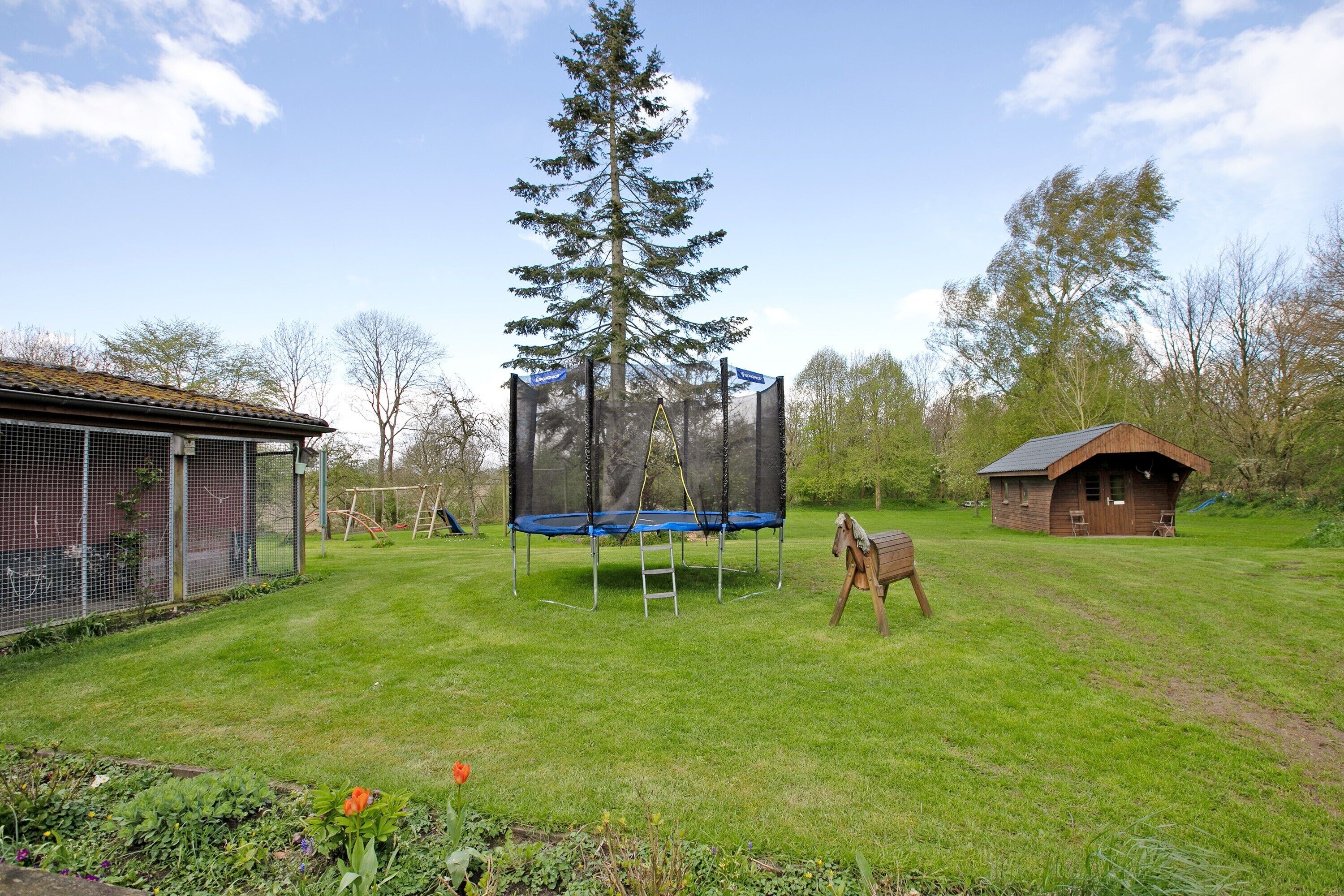 Een omheind gazon met een trampolaan, een houten paard en een klein houten gebouwtje.