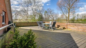 Eine Holzterrasse mit Tisch und Stühlen, ein Backsteinhaus und im Hintergrund kahl stehende Bäume unter einem teils bewölkten Himmel.