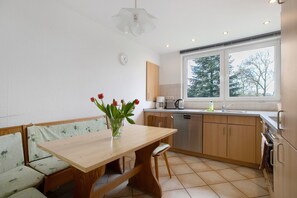 Una cocina con mesa de madera, asientos tipo banco, un ventanal con vista a árboles y un reloj en la pared.