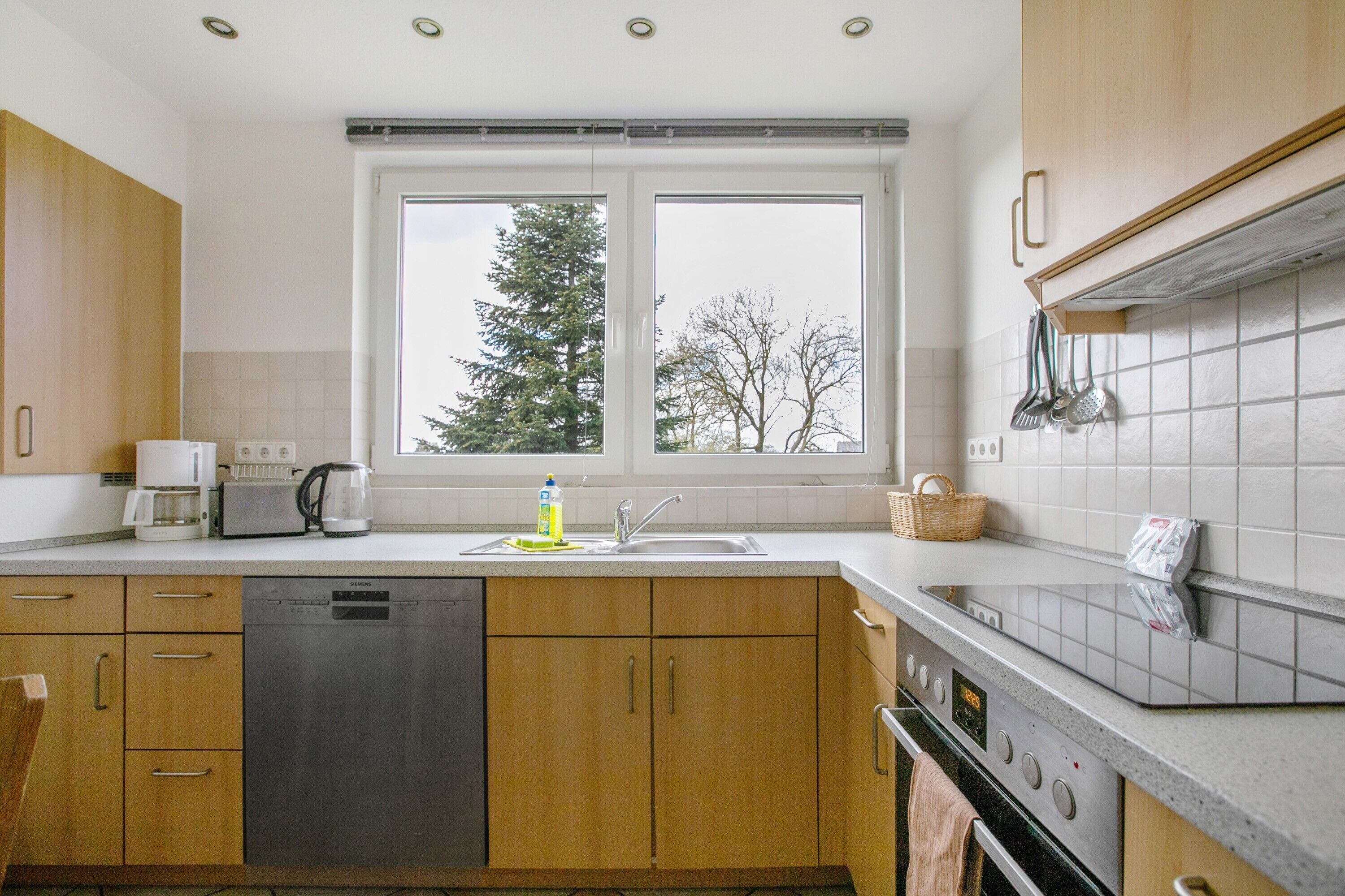 A modern kitchen with wooden cabinets, a dishwasher, a coffee maker, and a window with a view of trees.