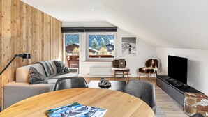 A modern living room with a wooden accent wall, a sofa with a blanket, a coffee table with magazines, a TV, and a view of a snowy mountain through large windows.