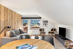 Un salon moderne avec un mur d’accent en bois, un canapé recouvert d’un plaid, une table basse avec des magazines, un téléviseur et une vue sur une montagne enneigée par de grandes fenêtres.