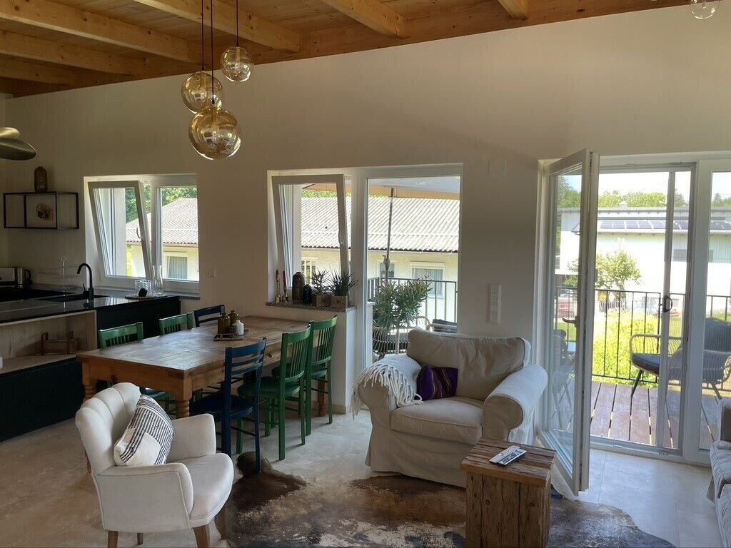 A spacious living area with a dining table, chairs, a sofa, and a coffee table. There are large windows providing natural light and views of the outdoors.
