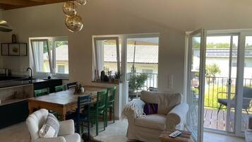 A spacious living area with a dining table, chairs, a sofa, and a coffee table. There are large windows providing natural light and views of the outdoors.