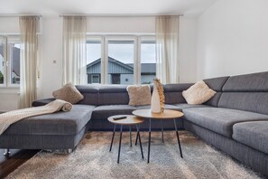 Un soggiorno moderno con un divano angolare grigio, un tavolino rotondo e vista su una casa attraverso ampie finestre.
