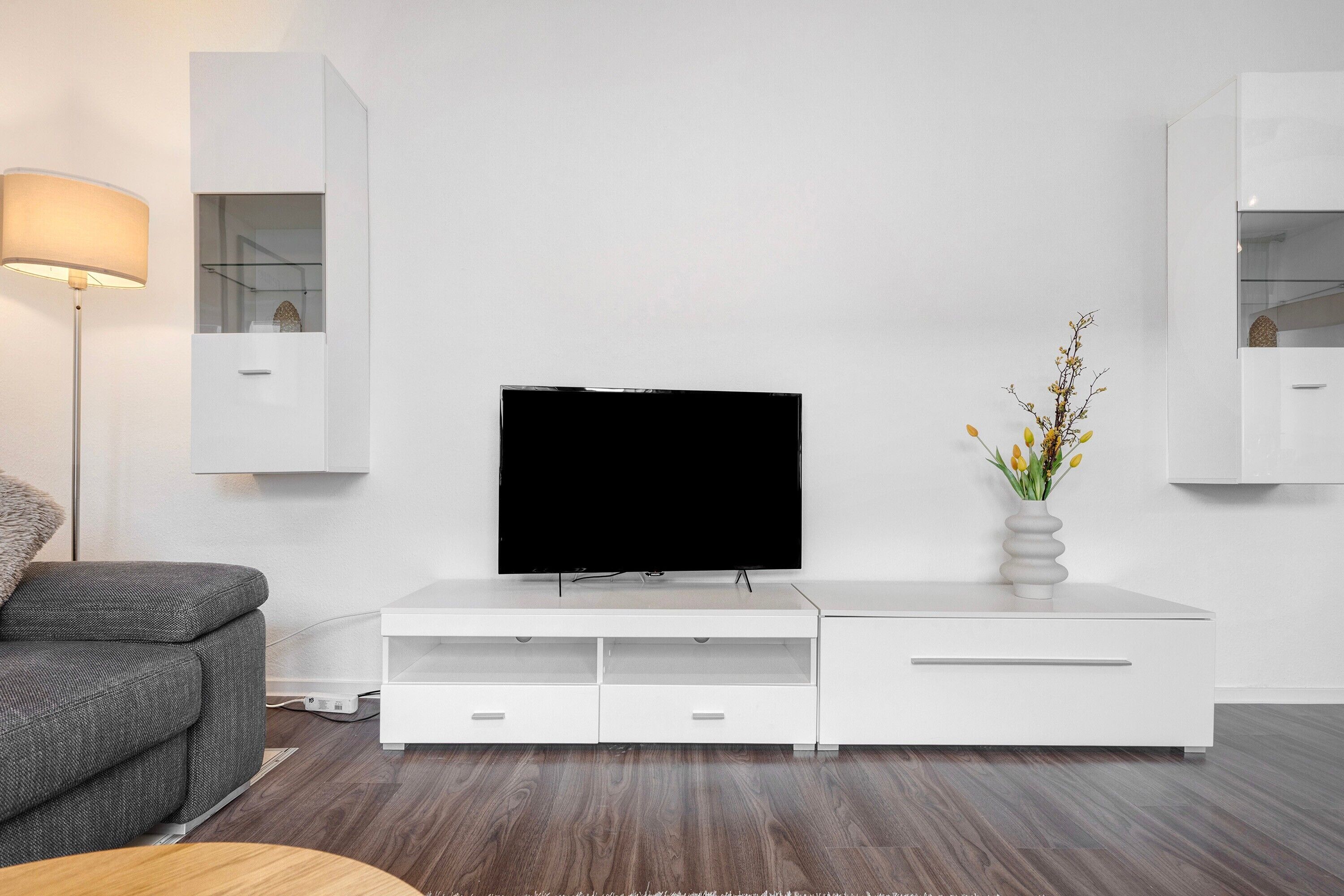 Ein modernes Wohnzimmer mit Flachbildfernseher, weißem Unterhaltungsschrank, grauem Sofa und einer Stehlampe.