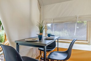 Un espace repas aménagé sous une tente, avec une table dressée pour deux personnes, deux chaises, une plante en pot et une fenêtre donnant sur un paysage verdoyant.