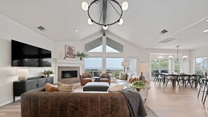 A spacious living room with a large sectional sofa, a fireplace, and a dining area with a high ceiling and large windows. - Private Hill Country Pool Retreat w/ Skyline Views (Canyon Lake)