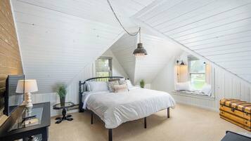 Une chambre avec un plafond en pente, un lit, une table de chevet avec une lampe, une fenêtre donnant sur un beau paysage et une commode en bois.
