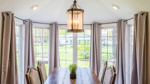 A dining area with a wooden table, six chairs, a hanging light fixture, and large windows with curtains. - Renovated Farmhouse w/ Game Barn & Firepit (Appleton)