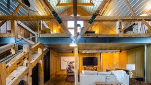 A spacious loft-style room with a high ceiling, exposed wooden beams, a large window, a fireplace, a sofa, a television, and a decorative wheel. - Renovated Farmhouse w/ Game Barn & Firepit (Appleton)