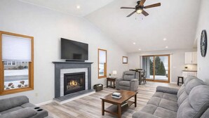 A spacious living room with a fireplace, a flat-screen TV, a ceiling fan, and a large gray sofa. - Stylish Townhouse w/ Patio & Free Parking (Appleton)