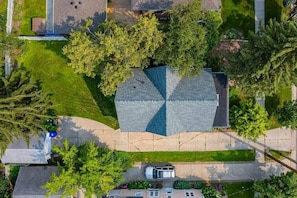 An aerial view of a residential area with a house featuring a unique geometric roof design, surrounded by lush greenery and a clear driveway.