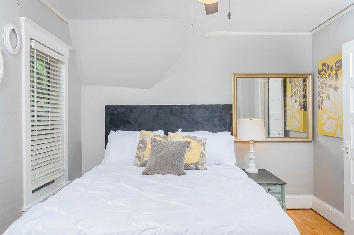 Een slaapkamer met een bed, kussens, een nachtkastje, een spiegel en een raam met jaloezieën.