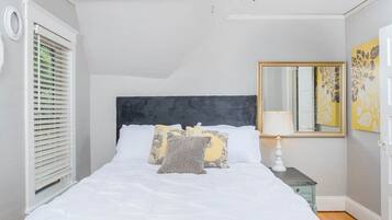 Een slaapkamer met een bed, kussens, een nachtkastje, een spiegel en een raam met jaloezieën.