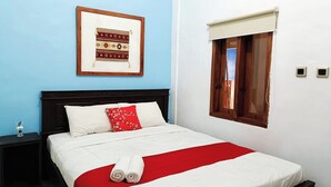 A neatly made bed with a red and white bedspread, a pillow with a red floral pattern, and two rolled towels. There is a framed picture on the wall above the bed. - Hotel O Ipeda Family Residence (Banyuwangi)