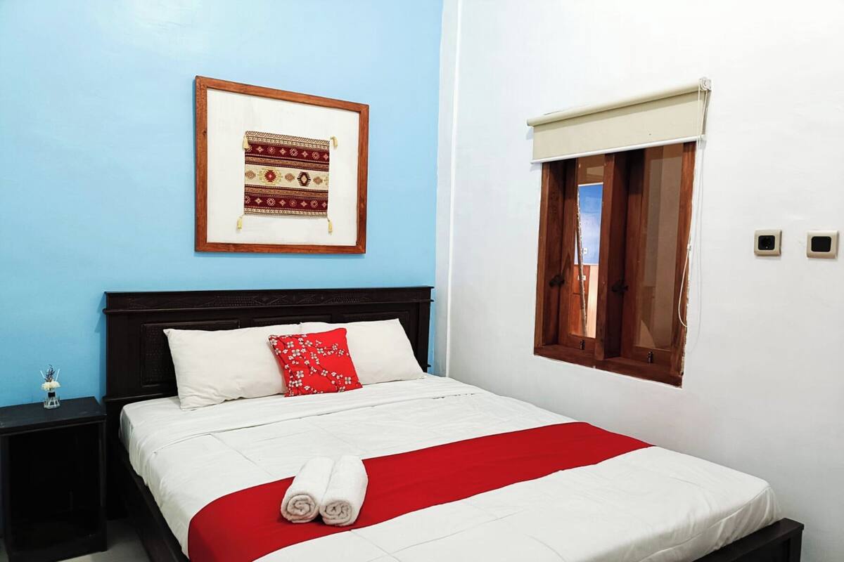 A neatly made bed with a red and white bedspread, a pillow with a red floral pattern, and two rolled towels. There is a framed picture on the wall above the bed.