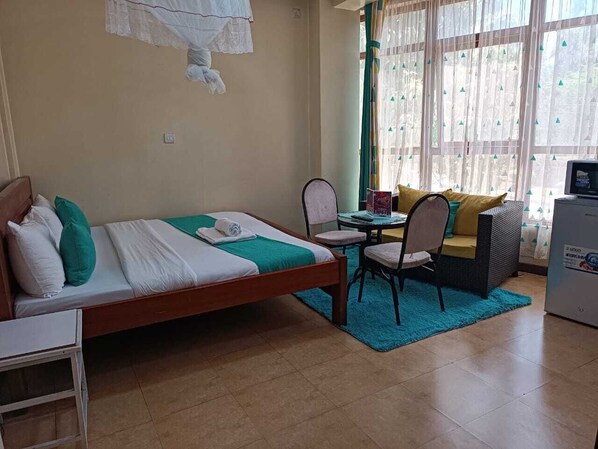 A compact hotel room with a single bed, a small table with a chair, a sofa, and a refrigerator.