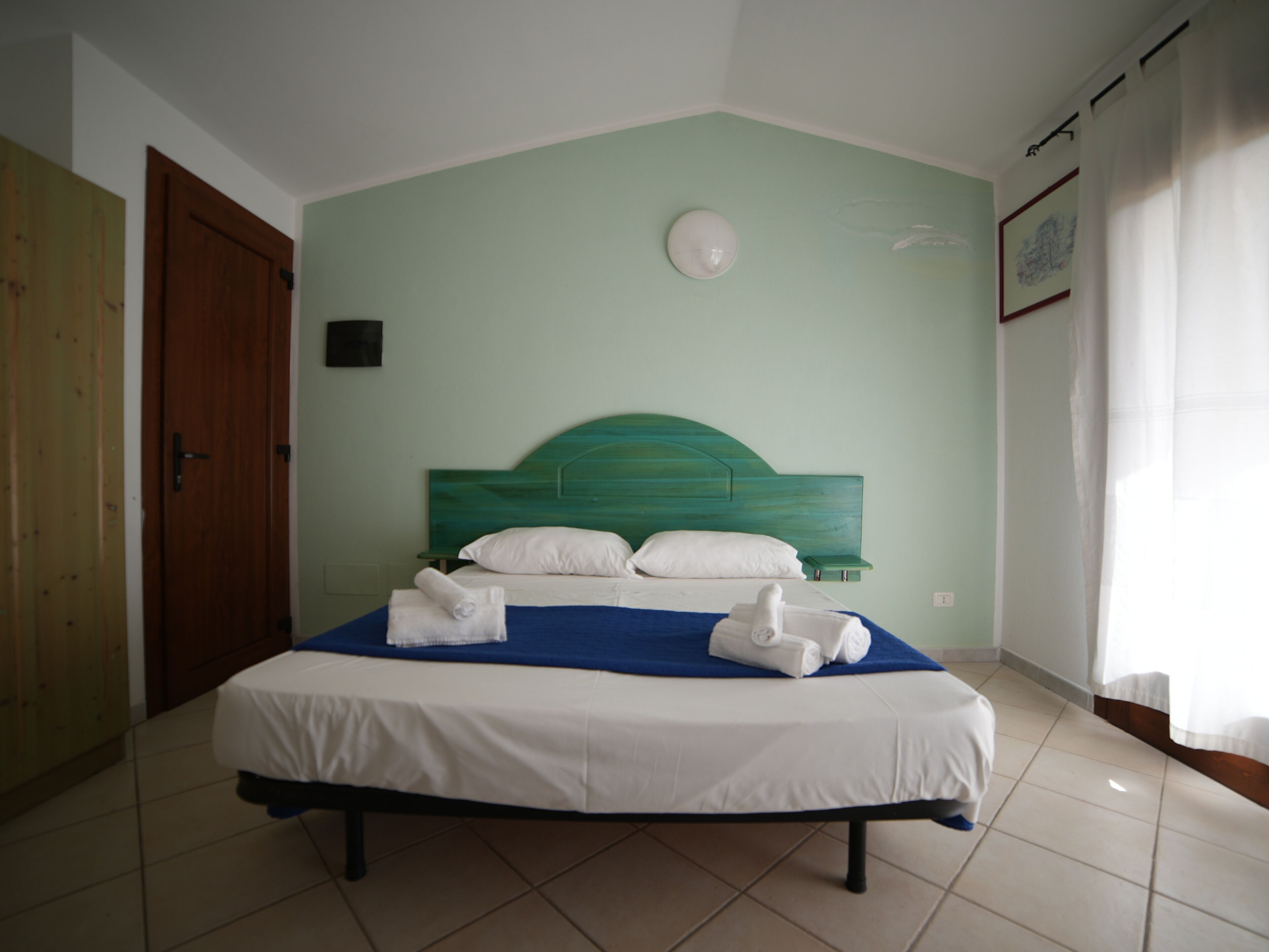 Un lit bien fait, avec du linge de lit blanc et un élément décoratif bleu, une porte en bois sur la gauche et un mur vert clair avec un luminaire rond au-dessus du lit.