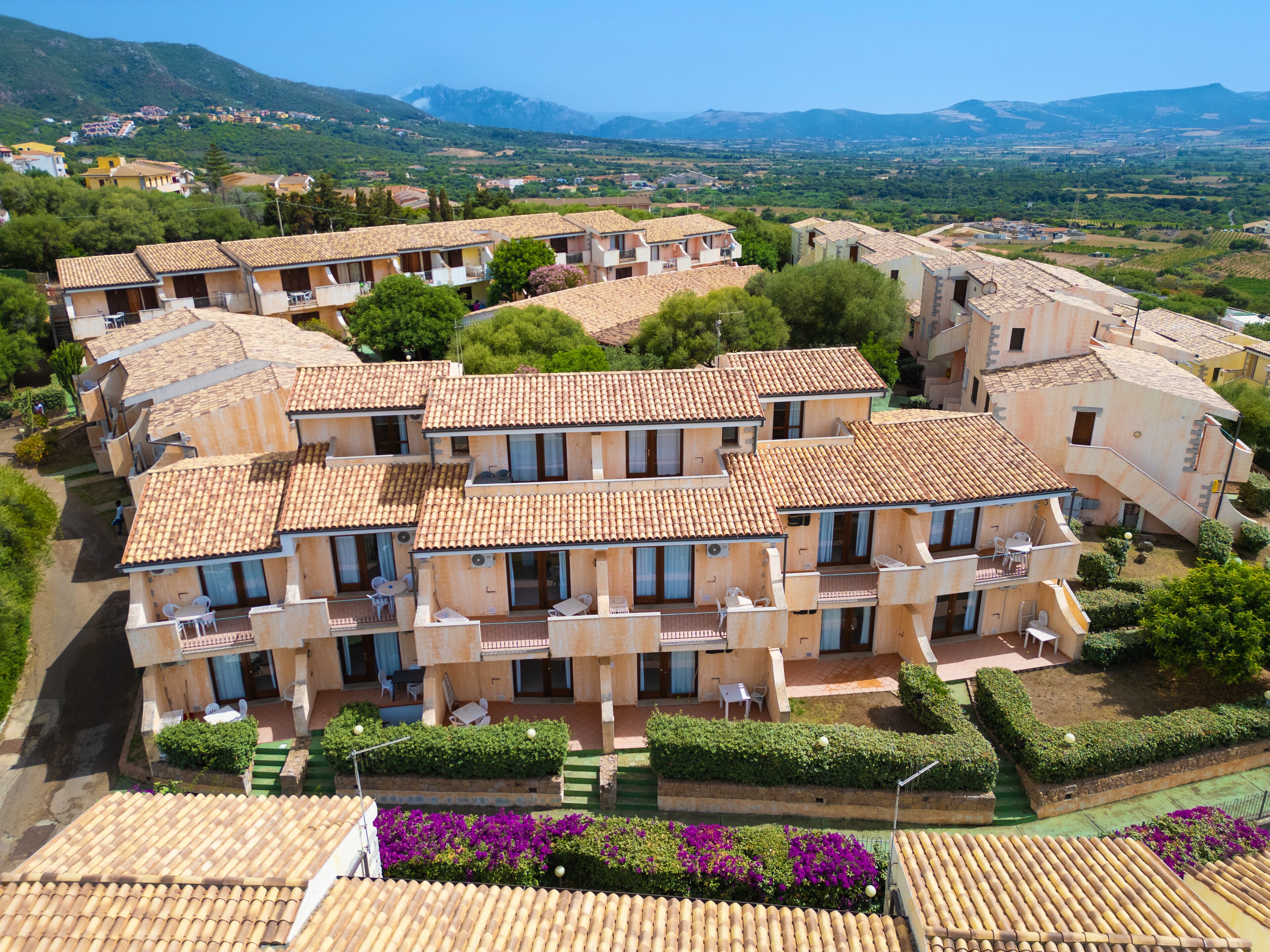 Un conjunto de edificios de estilo mediterráneo con techos de tejas de arcilla, organizados en un patio con jardines cuidados y vistas del paisaje circundante.