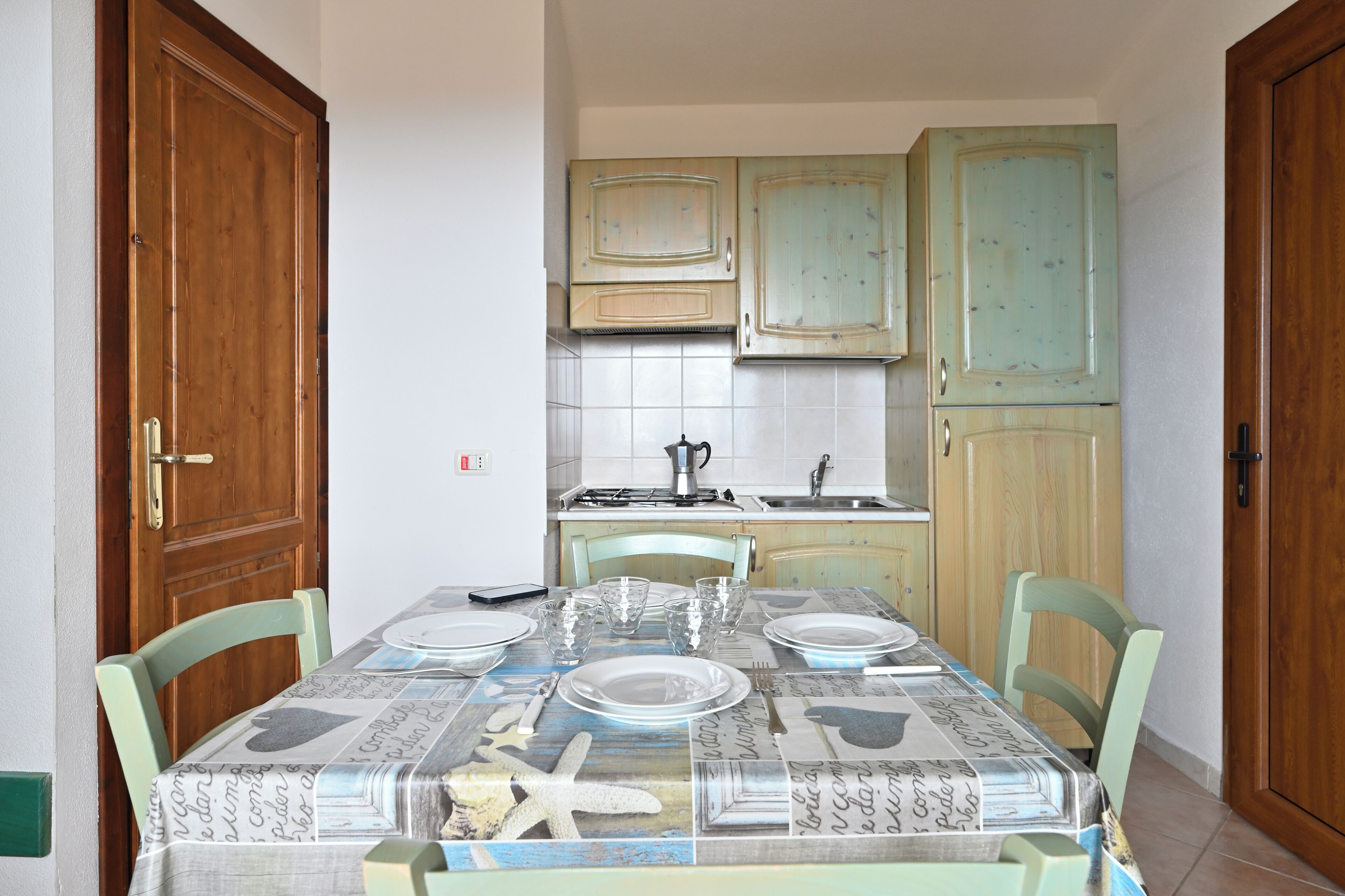 A dining area with a table set for four, surrounded by chairs. There is a kitchen area with cabinets and a stove in the background.